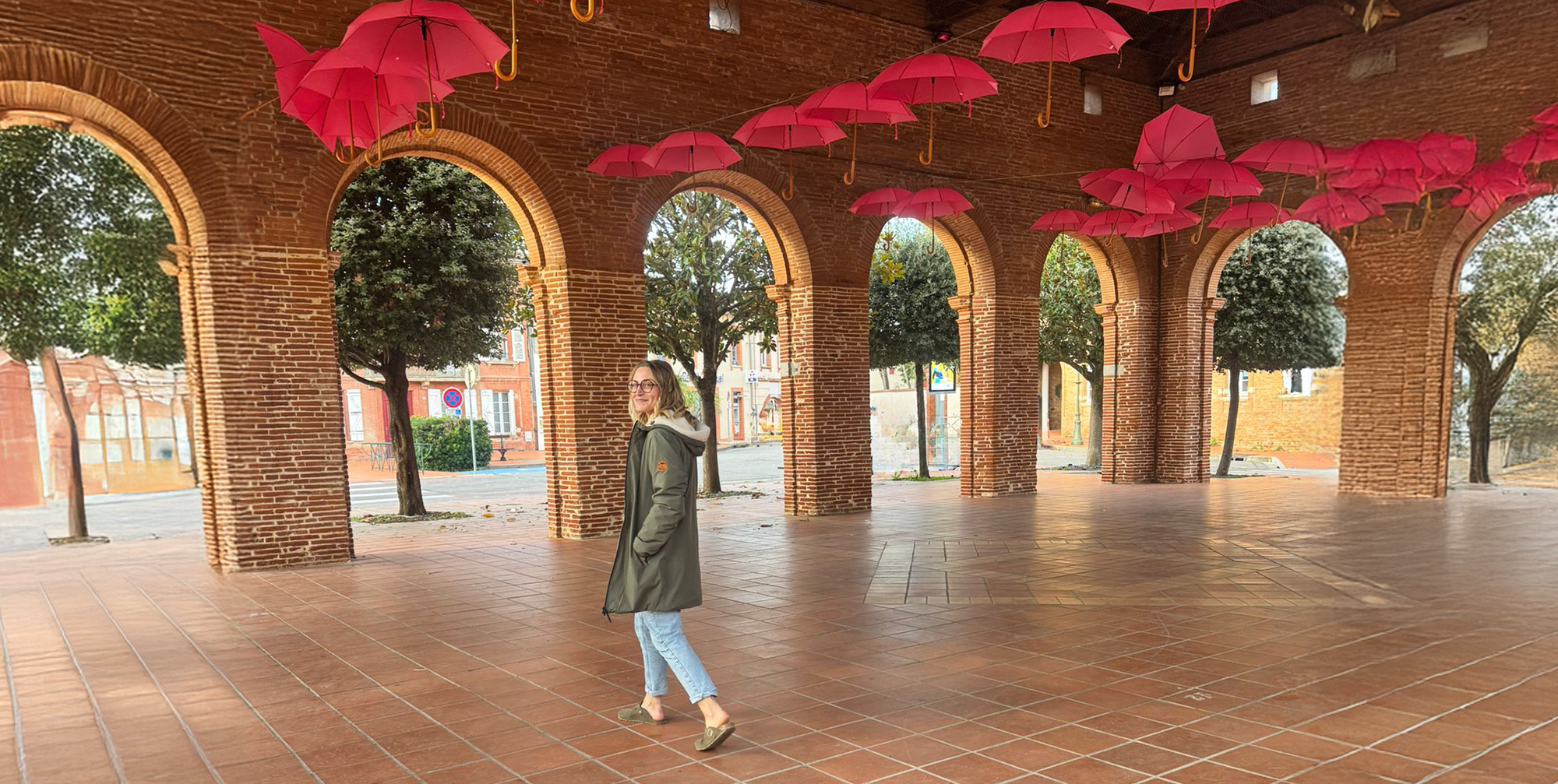 Femme en parka longue kaki doublé sherpa sous des arches en briques, esprit slow travel