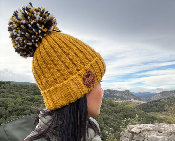 Détail du bonnet jaune avec pompon, porté face à un paysage montagneux.