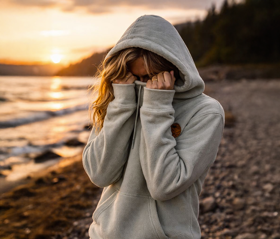 Femme cachant son visage avec le sweat polaire vanlife en bord de mer au couché du soleil