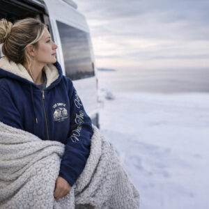 Femme adossée à son van aménagé lors d'un road trip en montagne sous la neige portant le sweat chaud vu de 3/4