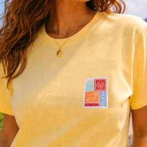 femme de face portant un tee shirt bio le tee shirt est jaune ananas le visuel retro fait penser à un timbre avec des motif surfeur / van life sur la poitrine